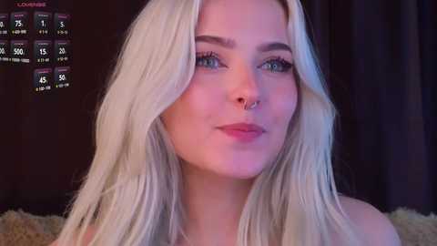 Video of a young, fair-skinned woman with long, platinum blonde hair, green eyes, and a septum piercing. She wears a subtle makeup look. Dark background with a calendar showing dates and temperatures.