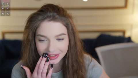 Video of a young Caucasian woman with long, wavy brown hair and fair skin, wearing a grey top, smiling while biting her black-painted fingernails. Background shows a beige wall and a dark blue sofa.