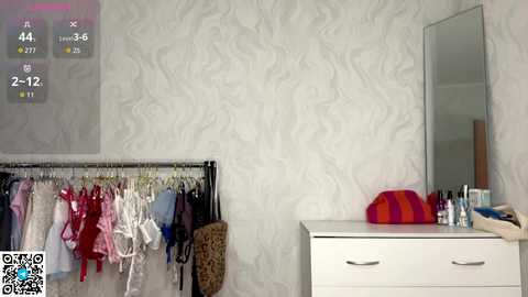 Video of a bedroom with a white, textured wall, a black clothing rack displaying various lingerie, a white dresser holding makeup and a red hat, and a large mirror.