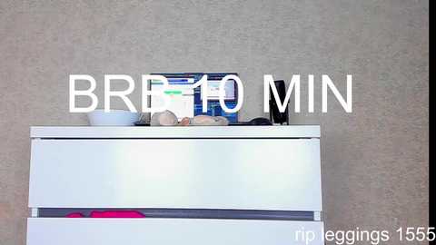 Video of a white dresser with a can of BRRR 10 MIN spray cleaner, a pink handbag, and a bowl on top, set against a textured beige wall.