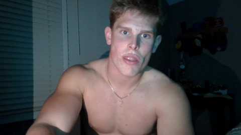 Video of a young, shirtless, fair-skinned man with short brown hair, wearing a gold chain necklace, in a dimly lit room with a messy background.