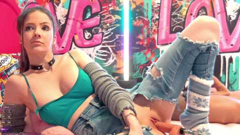 Video of a fair-skinned woman with dark hair, wearing a green bra and ripped jeans, lounging against a vibrant, graffiti-covered wall with the word \"VELVET\" in large, pink letters.