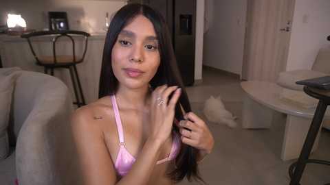 Video of a young woman with long, dark hair, wearing a pink bra, sitting in a modern living room with beige furniture, a white cat, and a kitchen in the background.