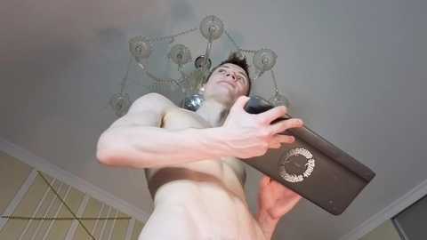 Video of a shirtless, fit young man with short brown hair, holding a black, circular, retro-style device, against a chandelier and beige walls.
