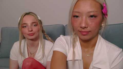Video of two young women with fair skin, blonde braided hair, and light pink lipstick. They wear white tops and sit on a blue sofa against a plain white wall, smiling softly.