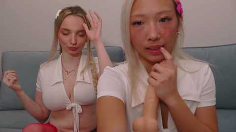 Video of two blonde women with fair skin, one in a white crop top, the other in a white shirt, both holding a large pink dildo.