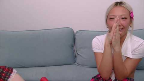 Video of an Asian woman with blonde hair, wearing a white top and plaid skirt, kneeling on a light blue couch, covering her face with her hands, smiling.