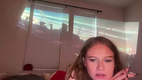 Video of a young Caucasian woman with long brown hair, wearing makeup, looking contemplative near a window with blinds.