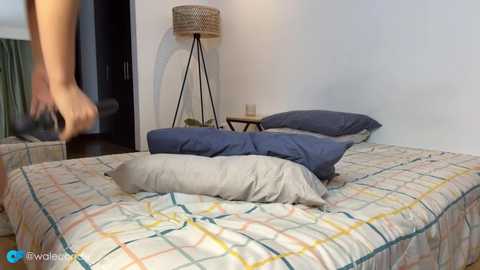 Video of a tidy bedroom with a beige and yellow checkered bedspread, a dark wooden floor, and a modern lamp on a small table.
