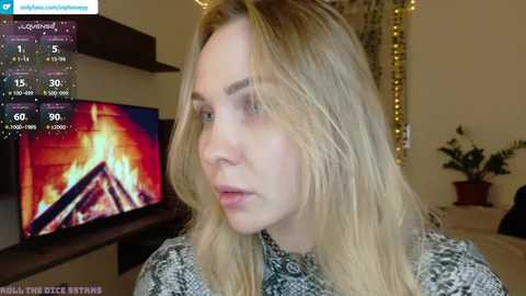 Video of a blonde woman in a patterned sweater, watching a TV with a fire screen. The background features a cozy room with fairy lights, potted plants, and a fireplace.