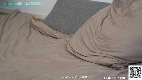 Video of a beige bed with wrinkled sheets, a grey pillow, and a grey blanket. The background includes a white wall and a light-colored floor. Text overlay includes a watermark and a QR code.