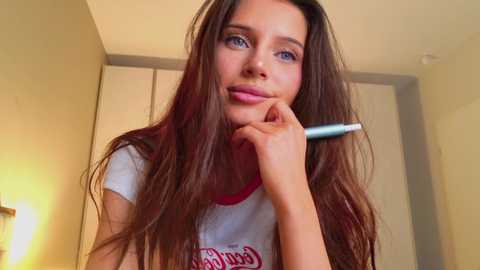 Video of a young woman with long brown hair, wearing a white t-shirt with red text, holding a blue pen, smiling in a beige-walled room.