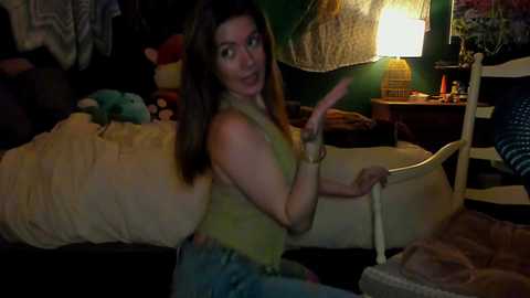 Video of a young woman with long brown hair, wearing a green tank top and jeans, sitting on a bed in a dimly lit, cozy bedroom.