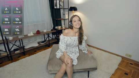 Video of a smiling Caucasian woman in a white off-shoulder dress, sitting on a beige cushion in a modern, minimalist living room with hardwood floors, white walls, and a black lamp.