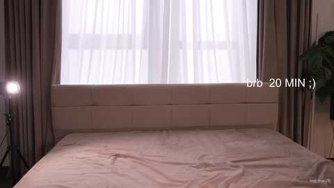 Video of a modern bedroom with a beige bedspread, sheer white curtains, and taupe floor-length drapes. A tripod lamp and a potted plant are visible on the left. Text: \"b6 20 MIN 3\" in the top right corner.