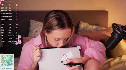 Video of a blonde woman in a pink blouse drawing a butterfly on a whiteboard with a black marker, set in a cozy bedroom with a warm, yellowish glow.