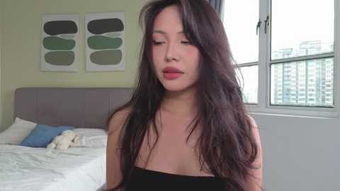 Video of an East Asian woman with long, wavy black hair, wearing a strapless black dress, standing in a modern bedroom with a bed, white walls, and a window.