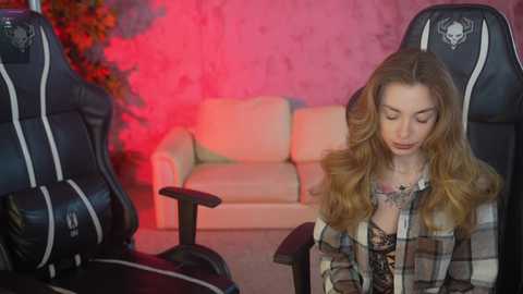 A video of a young woman with long, wavy blonde hair, wearing a plaid shirt and sitting in a gaming chair. The background features a cream-colored couch and a red-lit wall.
