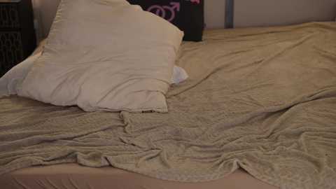 Video of a cozy bedroom with a beige, textured quilt on a bed, featuring a pillow and a partially visible person's arm. The background includes a dark dresser and a wall with a pink and purple floral pattern.