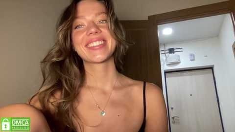 Video of a smiling Caucasian woman with wavy brown hair, wearing a black tank top, standing in a modern, sparsely furnished room with a large mirror and wooden door.
