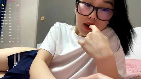 Video of an Asian woman with long black hair and glasses, lying on a bed, wearing a white t-shirt and navy blue shorts, biting her finger seductively.
