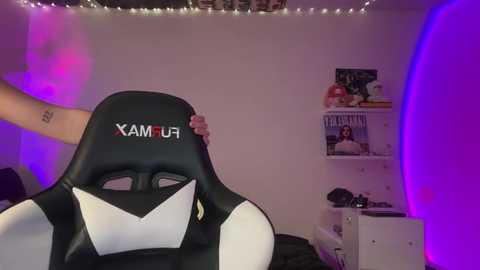 Video of a person sitting in a black and white \"XAMUR\" gaming chair, with a hand resting on the chair's headrest. The room features a white wall with purple LED lighting, shelves holding books and decor, and a plush, dark-colored couch.