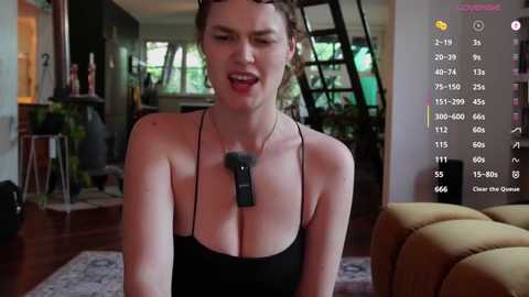 Video of a woman with fair skin, brown hair, wearing a black tank top and a facial massager device, in a modern, well-lit living room with greenery and wooden furniture.