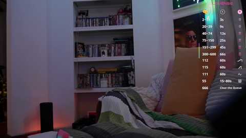 A video of a cozy bedroom with a man in a striped blanket, white bookshelves, and a TV displaying a chat window, set against a soft, ambient light.