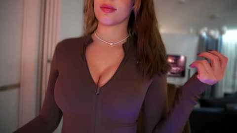 Video of a woman with light skin, long brown hair, and full lips, wearing a tight, dark brown zip-up top, standing indoors. Background shows a blurred room with a TV and modern decor.