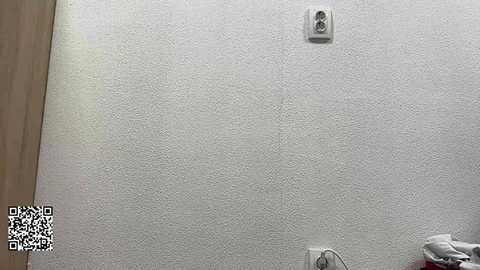 A video of a textured white wall with a single white electrical outlet near the center. The wall is slightly off-white with a subtle pattern. A QR code is in the bottom left corner.