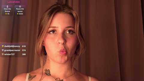 Video of a young woman with light skin and blonde hair, blowing a kiss, wearing a light-colored top. Background features dark brown curtains.