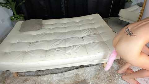 Video of a nude woman with a tattoo on her lower back, wearing a pink strap-on, standing next to a tufted cream leather chaise lounge in a modern, minimalistic room.