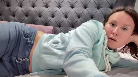 Video of a Caucasian woman with fair skin and brown hair lying on a bed, wearing a light blue hoodie and blue jeans, with a gray tufted headboard in the background.