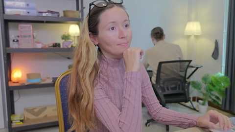 Video of a young woman with long blonde hair, wearing a pink sweater, seated at a desk in a modern office with shelves, plants, and two people working in the background.