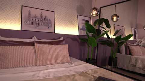 Video of a cozy bedroom with a bed adorned with beige and white striped pillows, surrounded by a lavender wall and two large potted plants. Framed artwork and a mirrored wall reflect the space.