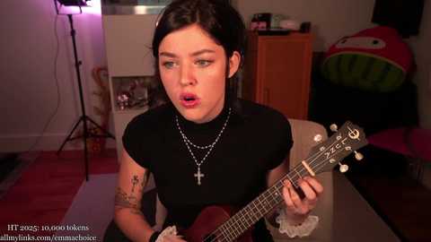 Video of a pale-skinned woman with short dark hair, wearing a black dress, playing an electric guitar, in a cozy living room with a \"Super Mario\" pillow and a studio light.