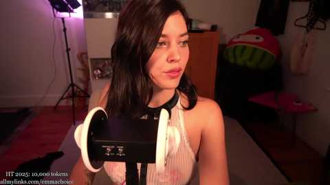 A video of a young woman with shoulder-length black hair, wearing a white sleeveless dress, holding a microphone in a brightly lit room with studio lighting.