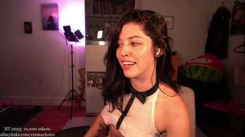A video of a smiling, dark-haired woman in a white ribbed top, wearing black headphones, sitting in a dimly lit room with studio lights and a Pikachu plushie.
