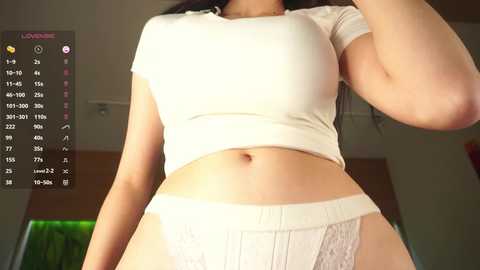 A video of a light-skinned woman with long dark hair, wearing a tight, white cropped top and lace panties, indoors with a green plant in the background.