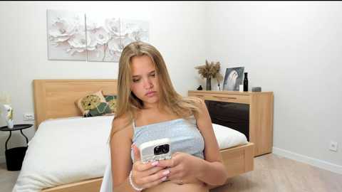 Video of a blonde young woman in a white camisole, sitting on a bed in a minimalist bedroom, holding a smartphone, with a wooden headboard and floral wall art.