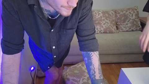 Video of a man with a beard and tattoos, wearing a black shirt, leaning over a beige couch with floral patterns, in a room with wooden flooring and a white coffee table.