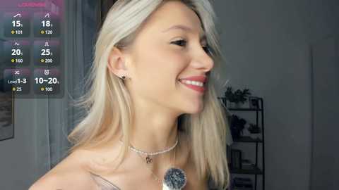 Video of a smiling, fair-skinned blonde woman with straight hair, wearing a silver necklace and earrings, set against a blurred indoor background.