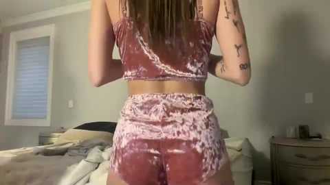 Media: Video of a woman with long brown hair, wearing a velvet pink and white tie-dye crop top and matching high-waisted shorts, seen from behind in a bedroom with a bed and window.