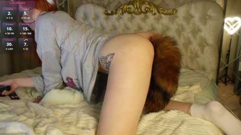 A video of a red-haired woman in a gray shirt, wearing a brown fur tail, on all fours on a beige bed, with a TV remote in hand.