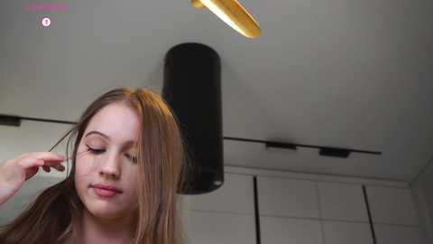 Video of a young woman with light skin and straight brown hair, wearing a white top, adjusting her hair in a modern, white-tiled room with a cylindrical black light fixture and recessed ceiling lights.