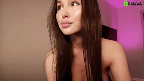 Media: A close-up video of a young, attractive woman with long, straight brown hair, fair skin, and a slender physique. She is topless, with visible cleavage, and has a contemplative expression. The background is a plain, beige wall with a pinkish glow.