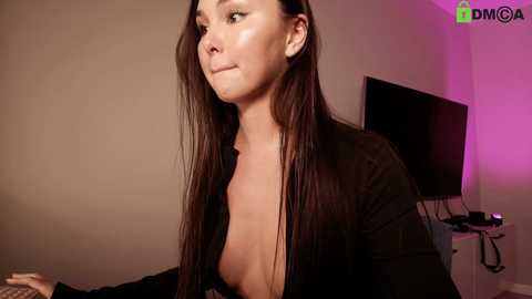 Video of a young woman with long dark hair, wearing a black cardigan and exposing her cleavage, in a dimly lit room with a TV and a purple light.