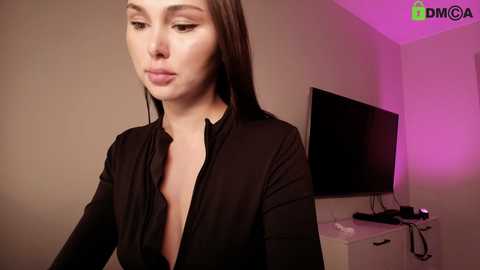 A video of a fair-skinned woman with straight, shoulder-length brown hair, wearing a black, unbuttoned blouse, in a room with a pink-painted wall, a flat-screen TV, and a white dresser.