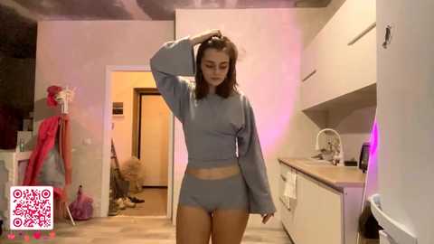 Video of a woman with long brown hair in a light blue crop top and matching shorts, standing in a modern kitchen with white cabinets and pink accents, holding her hair.