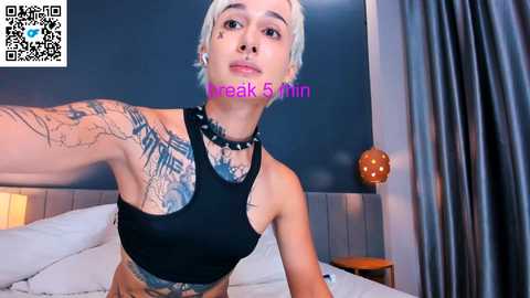 Video of a slender, light-skinned person with short platinum blonde hair, tattoos, and piercings, wearing a black crop top, sitting on a bed with white sheets.